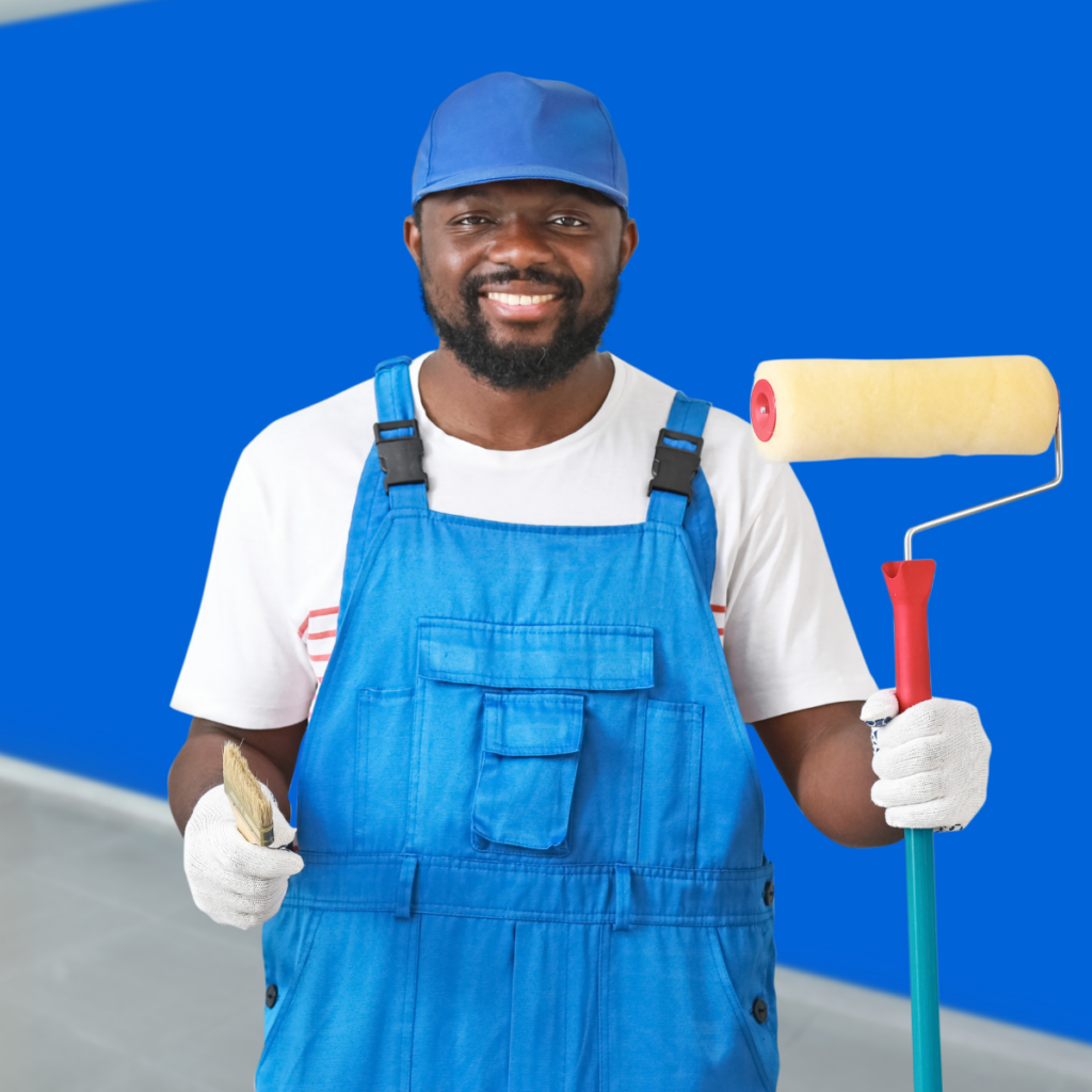 A man wearing painting overalls stands in front of a freshly painted blue wall. He holds a roller in one hand and a paintbrush in the other, ready to continue his work.