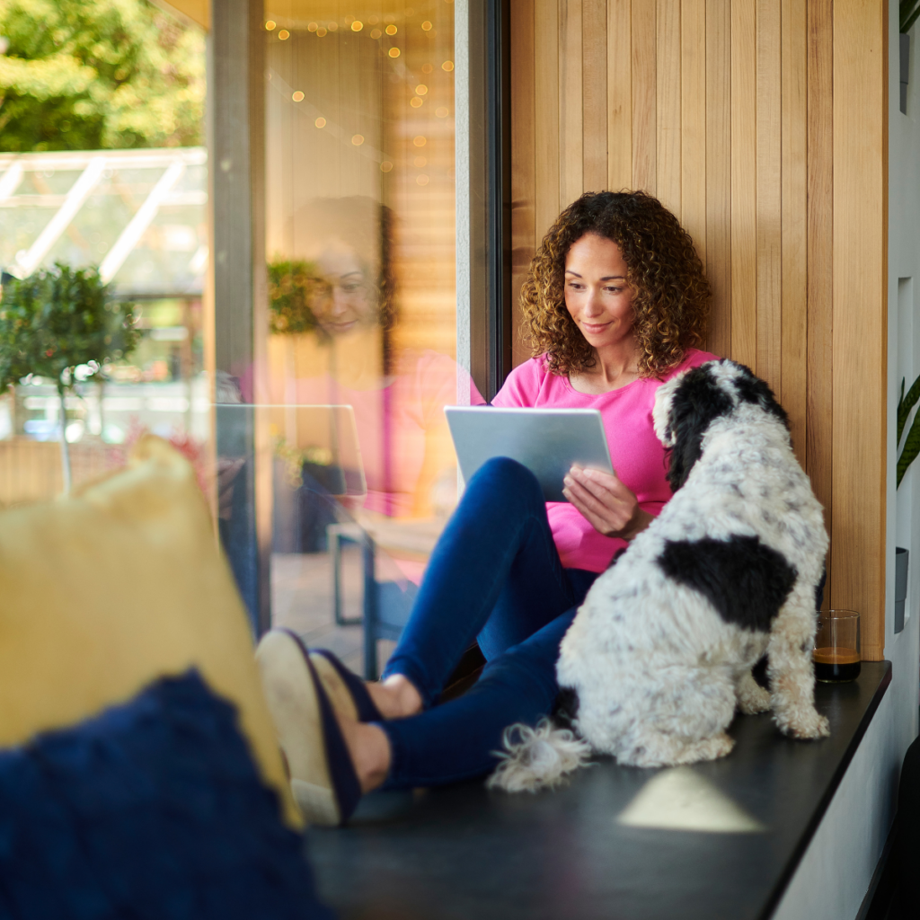Alt text: A woman sits on a cozy window bench with her legs crossed, engrossed in using a tablet. Her faithful dog sits beside her, both seemingly deep in thought. The woman appears focused, possibly reflecting on valuable information or conducting research relevant to her business.