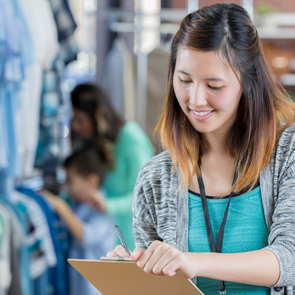 "Image of a focused young woman in a store, holding a clipboard and checking off tasks. Wearing a lanyard with work badge or keys around her neck. Customers can be seen in the background, browsing the items available in the store."