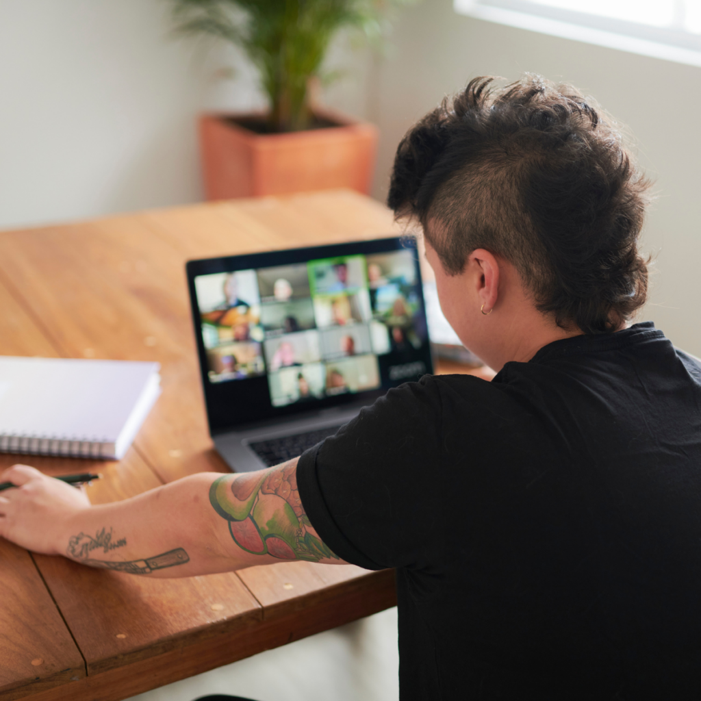 A person with an edgy appearance, including an unspoiled mohawk haircut, is sitting at a desk during a zoom call. They are discussing the financial aspects of their small business and taking notes in a notebook beside them.
