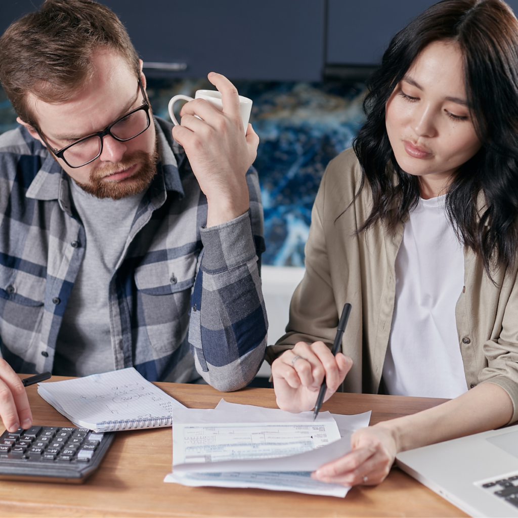 A woman and a man are seated at a table, engaged in a discussion about some paperwork. The woman, holding a pen, is pointing to specific sections of the documents as she explains the information to the man, who is attentively listening and looking at the papers. They both appear focused and collaborative, with notebooks and a laptop visible on the table.