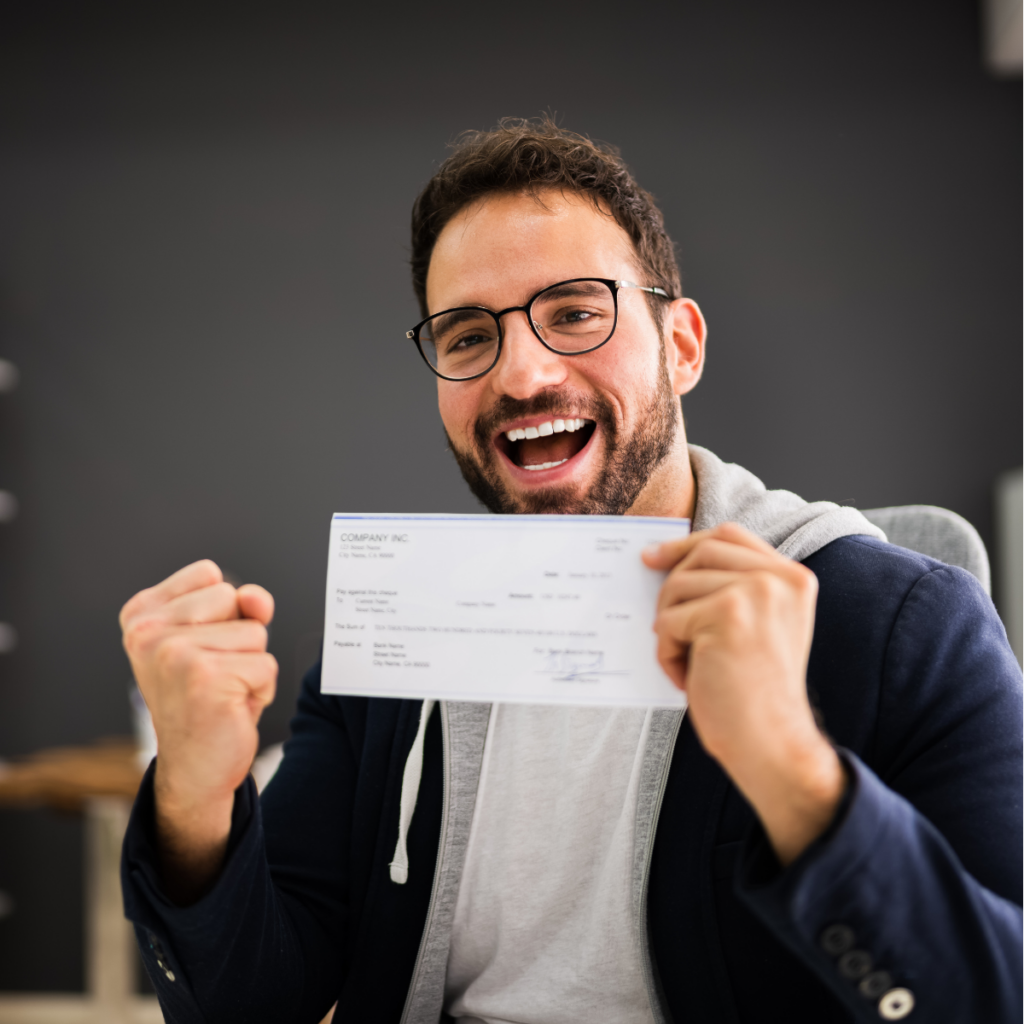 A cheerful young man sitting behind a desk, holding up a cheque with a proud smile on his face. He looks delighted, showcasing his accomplishment of writing the cheque or receiving it.