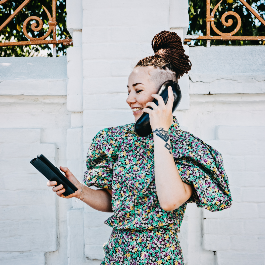 A smiling girl with a funky, trendy hairstyle is outdoors, holding a phone to her ear while engaged in conversation. Her vibrant style and expressive demeanor suggest that she is a creative individual or artist.