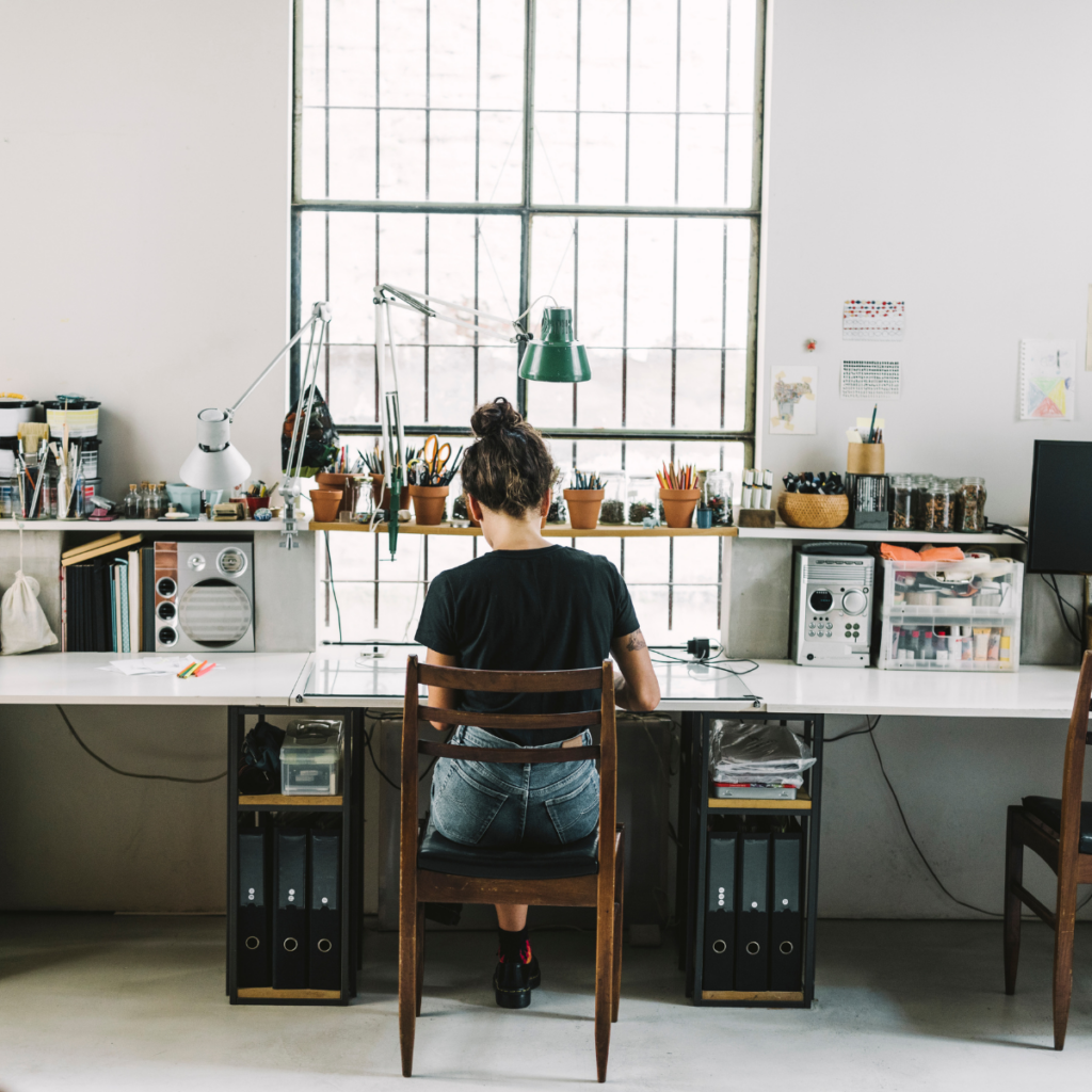 A woman with her back turned is seated at a long desk cluttered with paperwork and various creative materials. Shelves above the desk are also filled with documents and supplies. She is dressed in casual clothing, focused on her work.