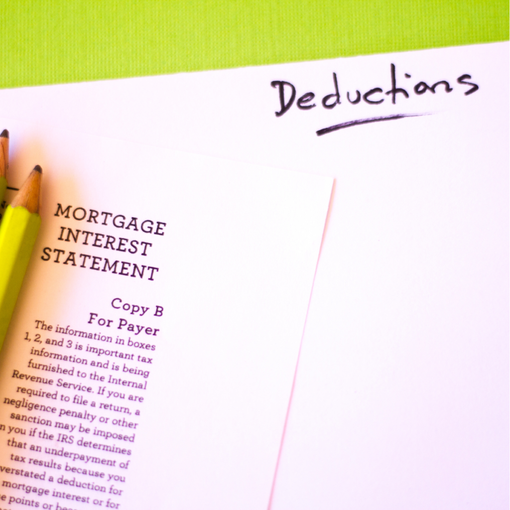 A tabletop with tax paperwork, two sharpened pencils, and a blank piece of paper. The top of the blank paper has "deductions" handwritten in neat cursive, suggesting that someone is preparing to make a list of tax deductions.