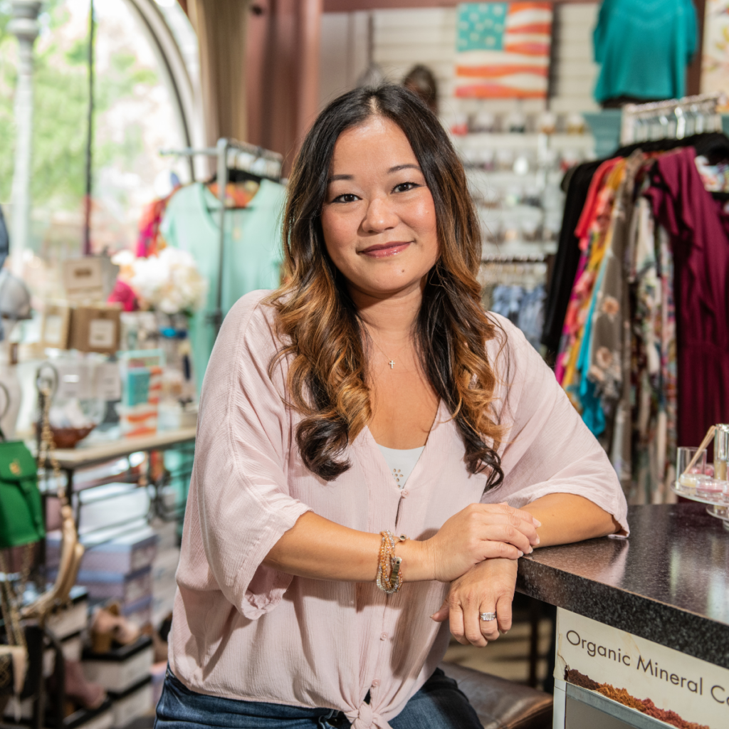 A middle-aged woman with a warm smile stands next to a counter in a vibrant shop filled with creative goods, including clothing and jewelry. The shop has a colorful and inviting atmosphere.