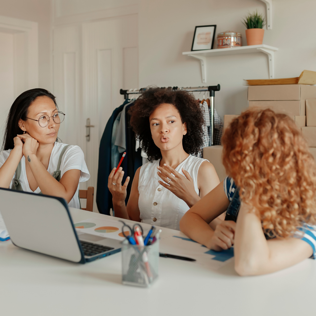 Three women collaborate at a large table desk in a creative loft office, surrounded by boxes on an adjacent desk. They are engaged in discussion, reviewing documents and exchanging ideas, with a dynamic and productive atmosphere evident in their expressions and body language.