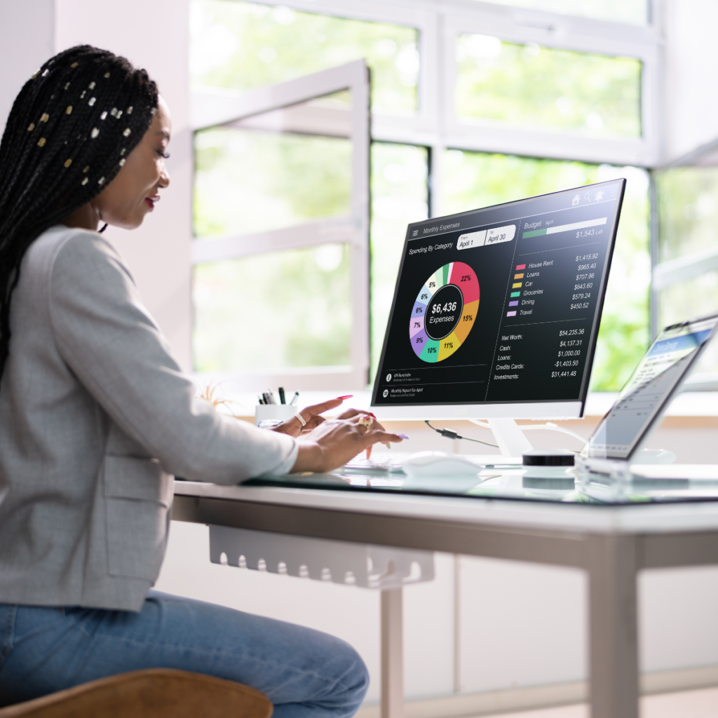 A young woman sits at a modern computer desk in a bright, open office, intently reviewing a large, colorful pie graph displayed on her screen. The workspace features minimalistic decor, with creative touches that enhance the airy atmosphere.