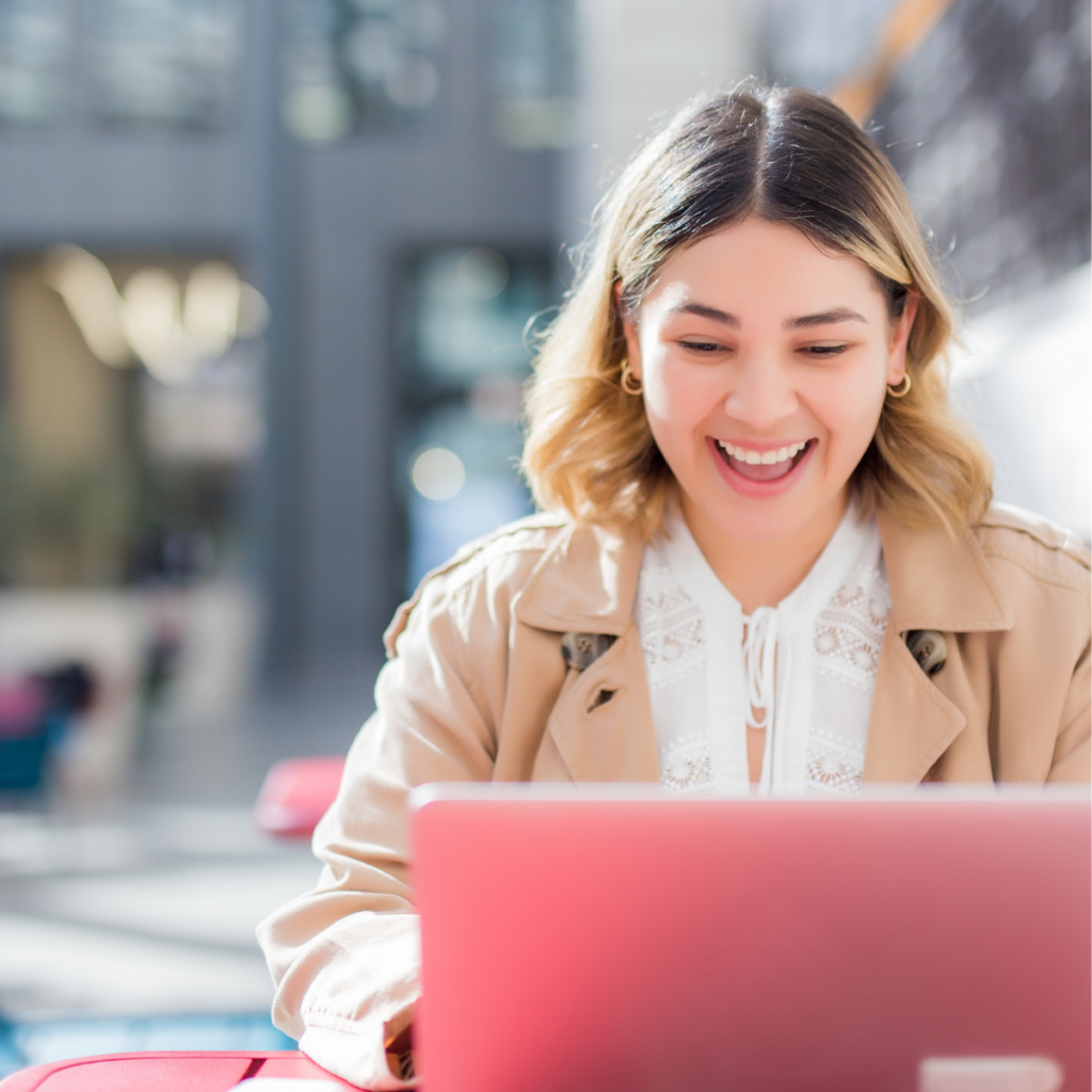 A young casual professional sits at an outdoor table, smiling while looking at her pink laptop. She is dressed comfortably and appears engaged and happy in her work environment. The background features sushine, suggesting a pleasant outdoor setting.