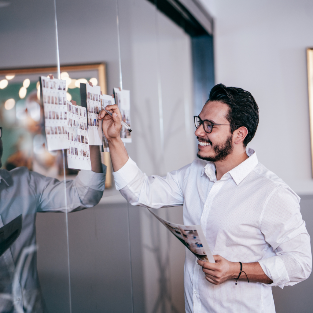 A man is focused on editing several pieces of paper taped to a mirror or window. The papers are filled with photographs, and he is using a pen to make notes or adjustments. The setting suggests a creative workspace, with natural light illuminating the scene.