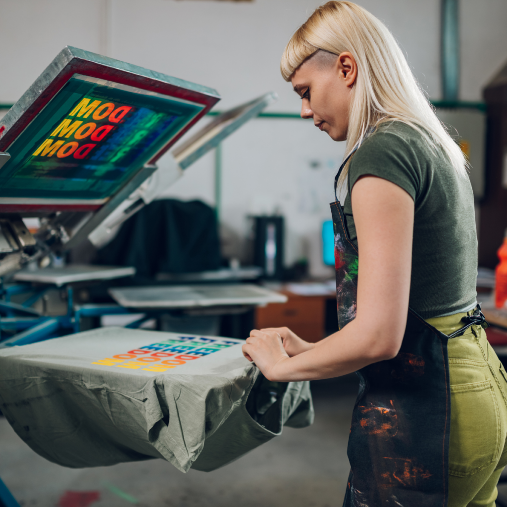 A woman with a funky style is using a t-shirt press to create graphic t-shirts in a spacious warehouse. The environment features large open spaces and various materials scattered around, showcasing her creative workspace.