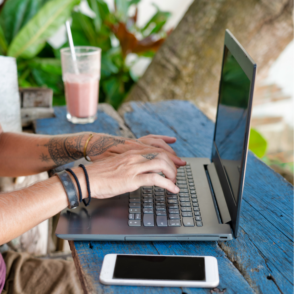Hands typing on a laptop placed on a table at a sunny beachside location, with sand, the ocean, and a tropical drink nearby