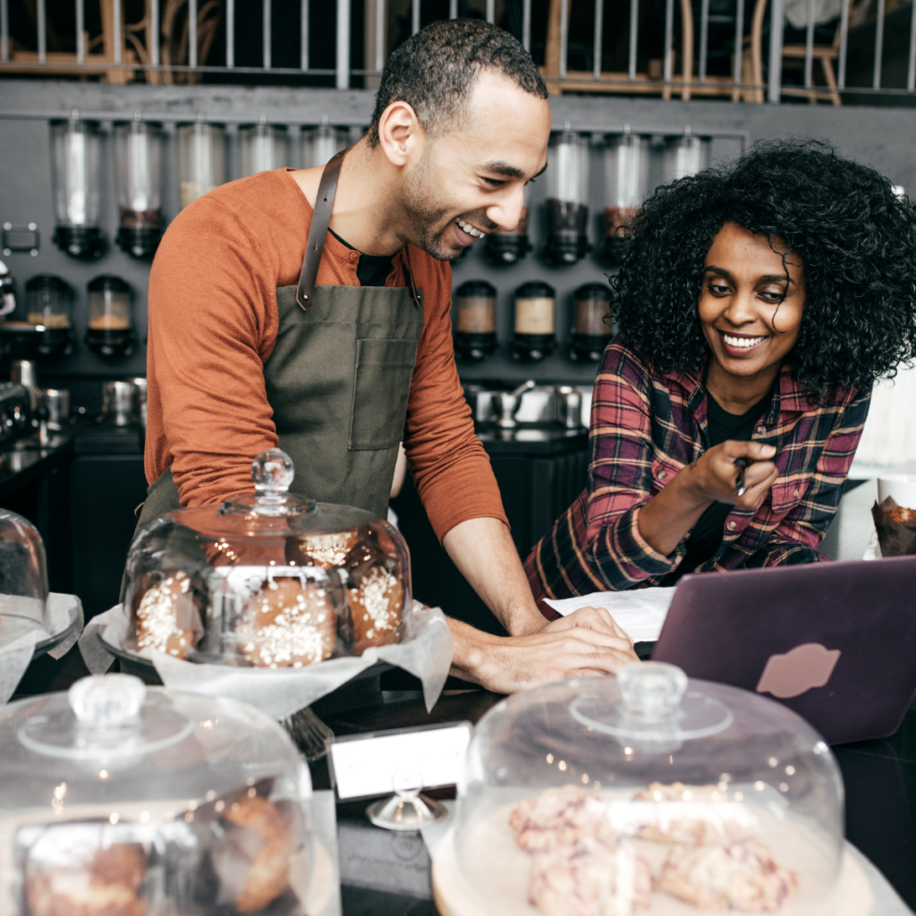 A man and woman stand behind a work table filled with baked goods and baking supplies. The man, wearing an apron, smiles as the woman looks at an open laptop displaying something on the screen. Both are smiling and appear engaged in their activity.