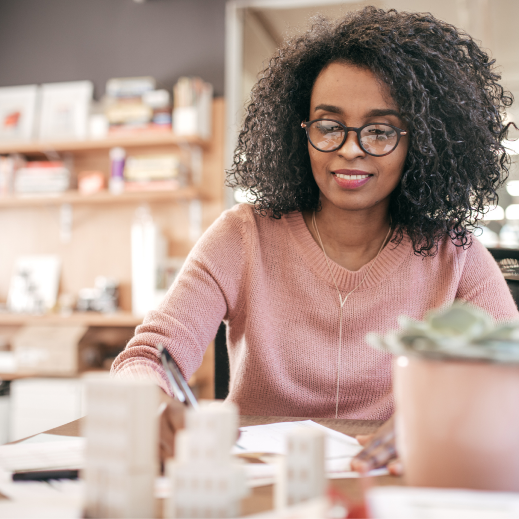 a middle aged woman sitting behind a desk in what appears to be a creative art studio. shes looking at something on the desk, perhaps paperwork.