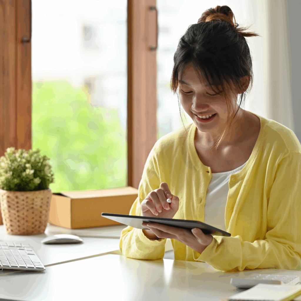 A young woman is sitting at a clean white desk next to a sunny window, smiling down at the screen of the tablet she is holding.