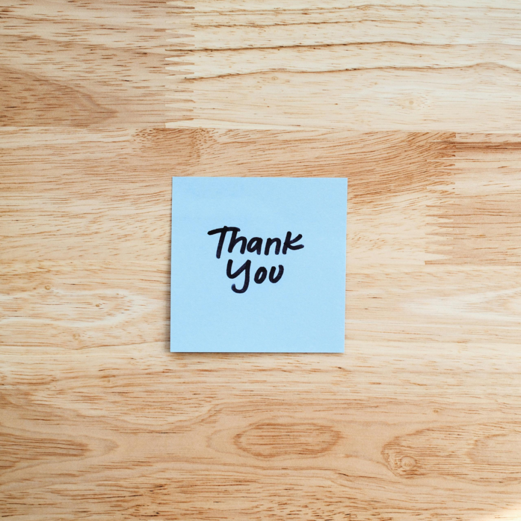 A minimalist overhead shot of a light-coloured wooden surface. A square blue post-it note sits alone in the middle, featuring a simple 