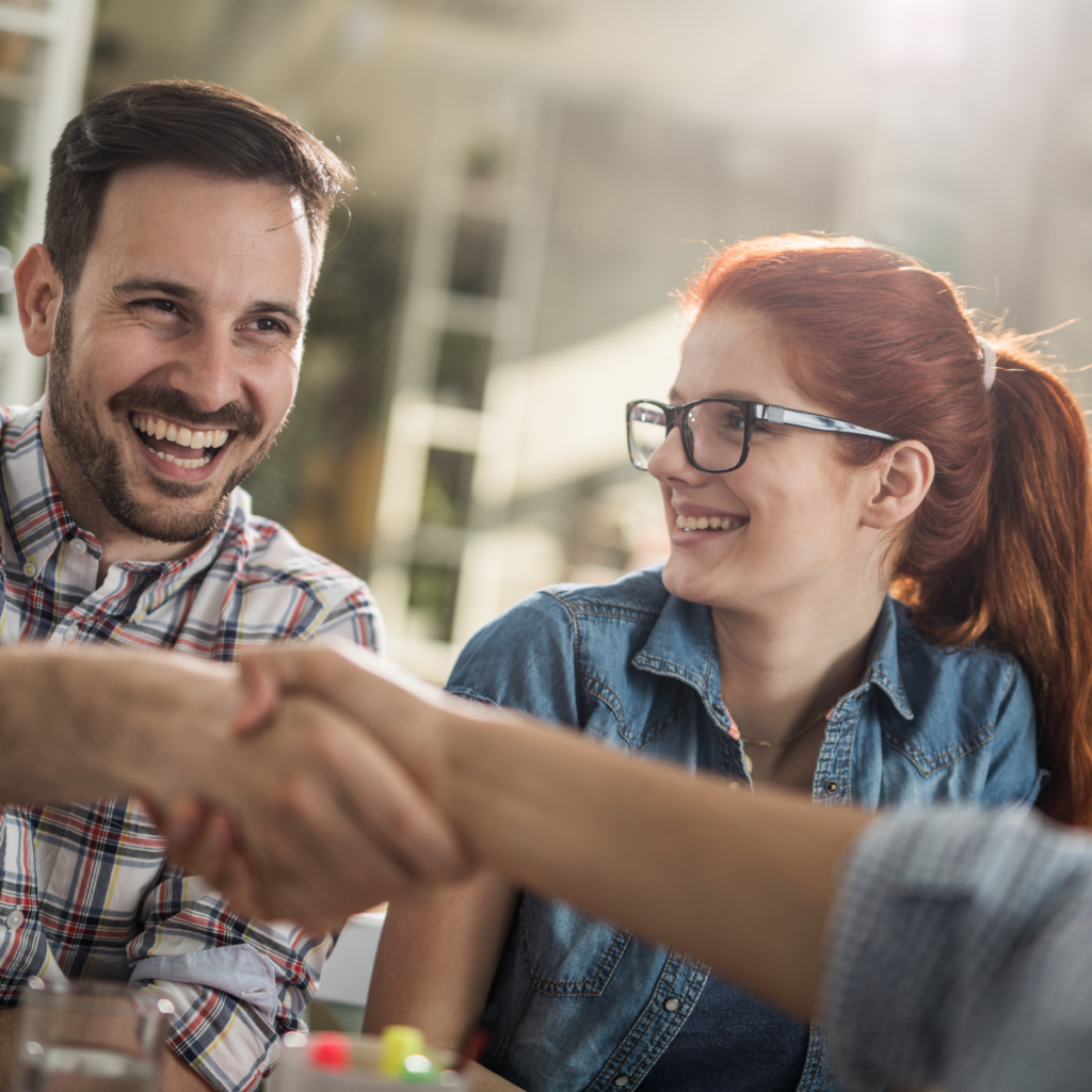 A straight-on shot of a man and a woman sitting side by side, both wearing bright, joyful smiles. The man is leaning slightly forward, extending his arm out of the frame to enthusiastically shake hands with an unseen person. The woman sits next to him, looking at him warmly with a proud and happy expression. The setting appears bright and professional, capturing a moment of mutual agreement or celebration.