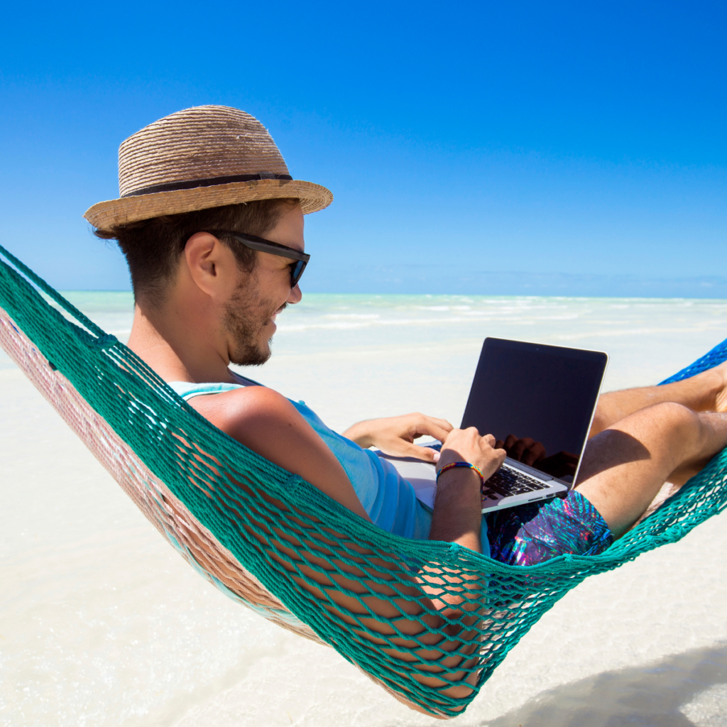 A person relaxed in a hammock suspended between palm trees on a tropical beach, working on a laptop resting on their lap. The individual is wearing a sun hat and facing away from the camera toward the bright, sunny ocean and sandy coastline. The scene is full of natural light and tropical details, with the man's face obscured by his pose and hat.