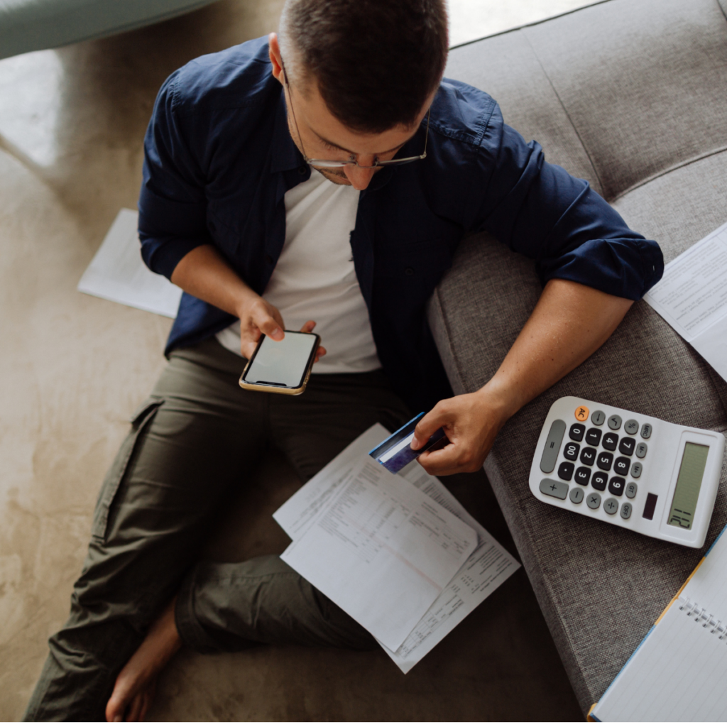 High-angle shot of a young man sitting on the floor leaning against a couch, comparing a credit card to paperwork and a phone while managing expenses.